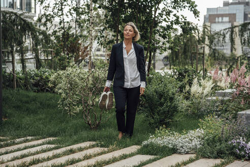 Female professional holding shoes while standing on grass at office park - VPIF04476