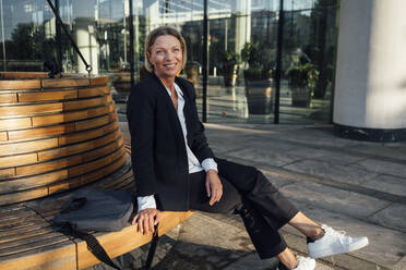 Smiling businesswoman with bag looking away while sitting at office park - VPIF04499