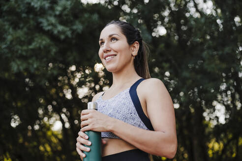 Lächelnde Sportlerin schaut weg, während sie eine Wasserflasche im Wald hält - EBBF04514