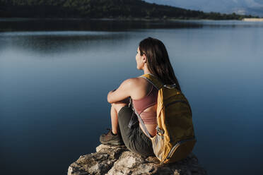 Junge Rucksackträgerin mit Rucksack auf einem Felsen am See sitzend - EBBF04520