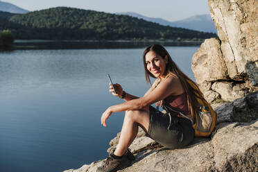 Lächelnde Wanderin mit Handy in der Hand auf einem Felsen am See sitzend - EBBF04521