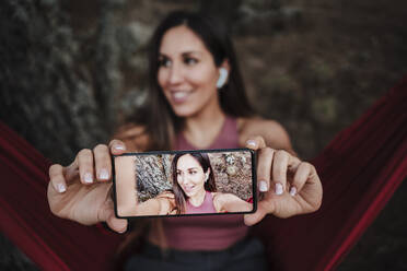 Lächelnde junge Frau nimmt Selfie durch Smartphone auf Hängematte - EBBF04529