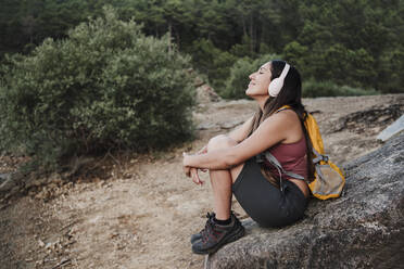 Weiblicher Rucksacktourist mit geschlossenen Augen, der auf einem Felsen sitzend Musik hört - EBBF04534