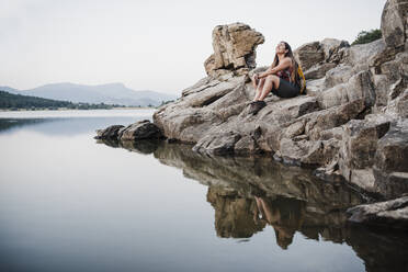 Smiling young woman with backpack looking up while sitting on rock near lake - EBBF04538