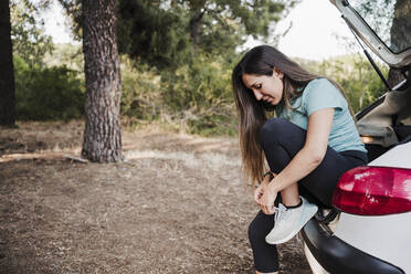 Young woman tying shoelace while sitting in car trunk - EBBF04549