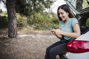 Lächelnde junge Frau, die ein Smartphone benutzt, während sie im Kofferraum eines Autos sitzt - EBBF04550