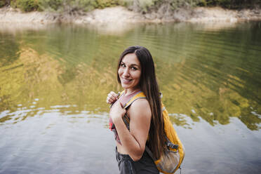 Smiling female backpacker standing by lake during vacation - EBBF04556