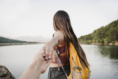 Female backpacker holding hand of friend at lakeshore - EBBF04560