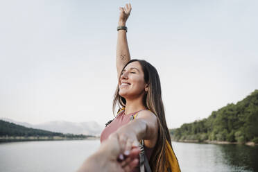 Carefree female hiker with eyes closed holding hand of friend by lake - EBBF04562