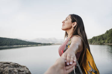 Female hiker with eyes closed holding hand of friend by lake during vacation - EBBF04563