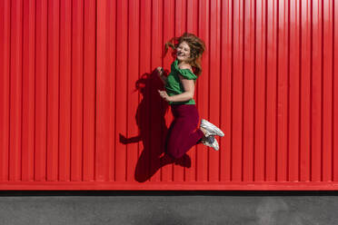 Fröhliche junge Frau springt durch rote Wellblechwand - VPIF04503