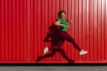 Young woman jumping on footpath by red wall during sunny day - VPIF04504