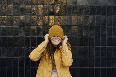 Verspielte junge Frau, die eine gelbe Strickmütze vor einer schwarzen Wand zieht - VPIF04530