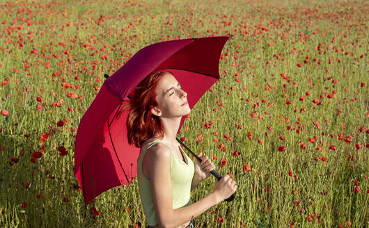 Rothaariges Mädchen mit geschlossenen Augen hält Regenschirm in Mohnfeld an sonnigem Tag - BFRF02363