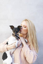 Young blond woman kissing dog in front of wall - EGHF00143
