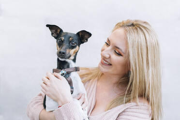 Smiling young blond woman looking at dog in front of wall - EGHF00149