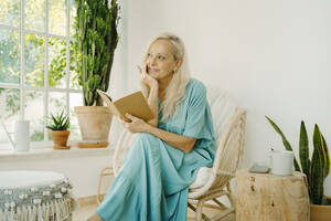 Thoughtful senior woman with hand on chin holding book while looking away at home - ERRF04931