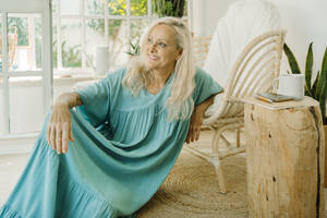 Smiling senior woman day dreaming while leaning on chair at home - ERRF04937