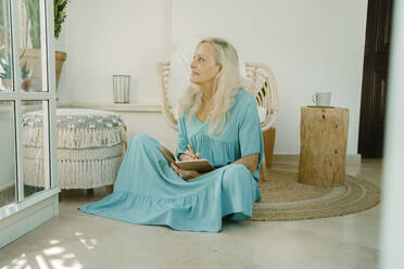 Thoughtful woman sitting with book on carpet at home - ERRF04939