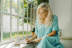 Senior woman with white hair writing in book near window at home - ERRF04943