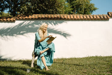 Woman reading book while sitting at backyard on sunny day - ERRF04962