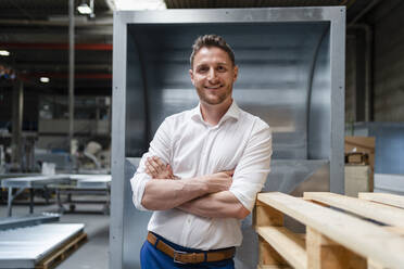 Smiling businessman with arms crossed leaning on wooden pallet - DIGF16168