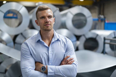 Confident young businessman looking away while standing with arms crossed at factory - DIGF16192