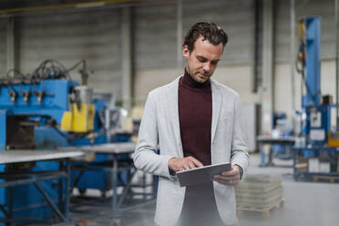 Männlicher Berufstätiger mit grauem Blazer, der bei der Arbeit in der Industrie ein digitales Tablet benutzt - DIGF16248