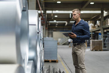 Young male inspector checking rolled sheet metal with digital tablet at warehouse - DIGF16278