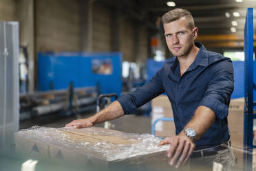 Confident young businessman staring while standing by cardboard box in industry - DIGF16296