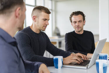 Junger männlicher Berufstätiger, der einen Laptop benutzt, während er mit Kollegen im Büro arbeitet - DIGF16300