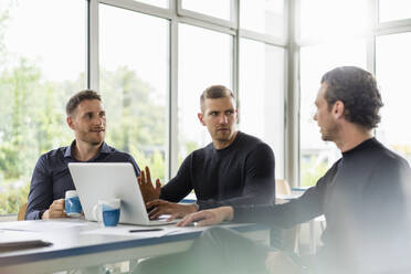 Male professionals sitting with laptop while discussing at desk in office - DIGF16301