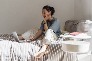 Female professional using laptop while sitting with hand on chin in bedroom - MEUF03719