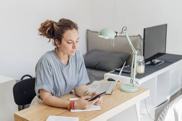 Female freelancer using mobile phone while sitting with laptop at apartment - MEUF03727