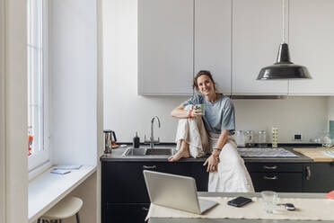 Lächelnde Freiberuflerin, die eine Kaffeetasse hält, während sie auf dem Küchentisch sitzt - MEUF03731