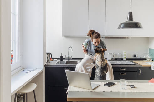 Weibliche Fachkraft spielt mit Hund, während sie in der Kaffeepause auf dem Küchentisch sitzt - MEUF03732