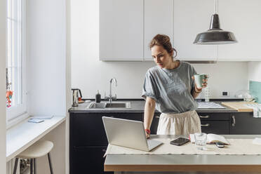 Woman holding coffee cup while using laptop in kitchen - MEUF03734