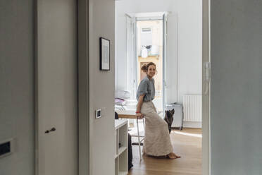 Smiling woman sitting on table looking through doorway in apartment - MEUF03739