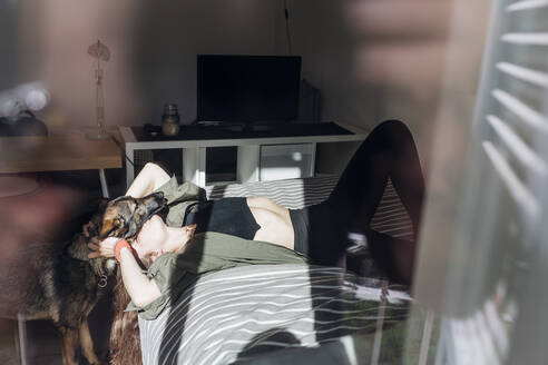 Woman kissing German shepherd dog while lying on bed seen from window - MEUF03743