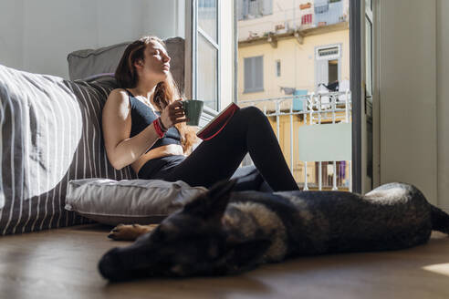 Frau mit Kaffeetasse und Buch entspannt sich bei ihrem Hund zu Hause - MEUF03753