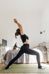 Woman with hands raised exercising on mat in bedroom - MEUF03758