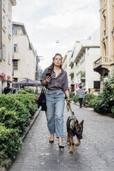 Frau hält Handy in der Hand, während sie mit ihrem Hund auf dem Gehweg spazieren geht - MEUF03777