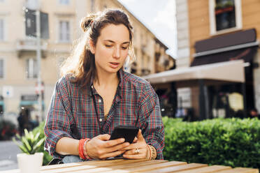Frau, die ein Smartphone benutzt, während sie an einem sonnigen Tag in einem Straßencafé sitzt - MEUF03779