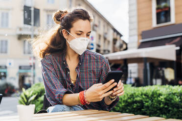 Woman with protective face mask using mobile phone at sidewalk cafe on sunny day - MEUF03780