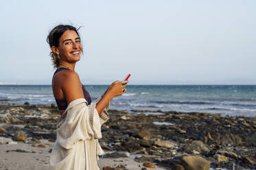 Glückliche junge Frau hält Handy am Strand - PGF00711