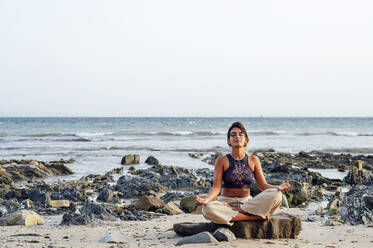 Young woman practicing lotus position while sitting on rock at beach - PGF00713