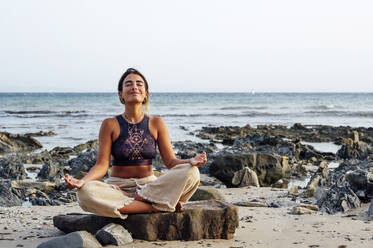 Smiling woman meditating on rock at beach - PGF00715