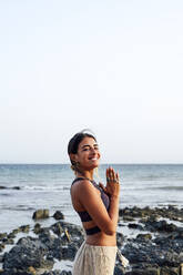 Smiling woman with hands clasped standing at beach - PGF00718