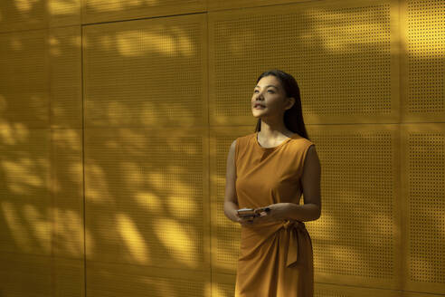 Beautiful young woman standing in front of yellow wall holding smartphone - EAF00034