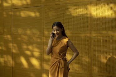 Smiling young woman wearing orange dress talking on mobile phone in front of yellow wall - EAF00036